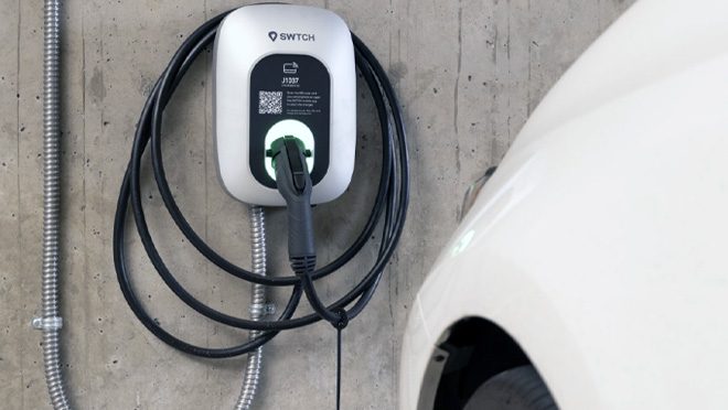 A wall mounted Level 2 EV charger on a concrete garage wall, cord coiled and the connector plugged into a parked car.